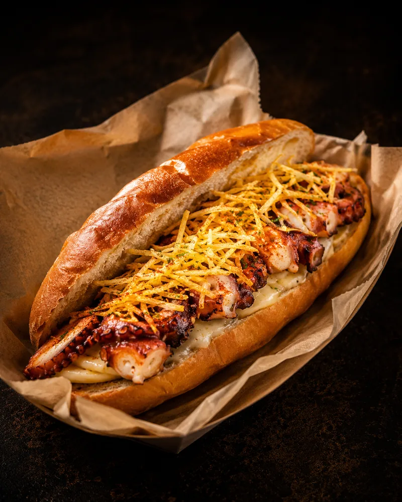 Rabodillo de pulpo: bocadillo largo en pan brioche con salsa blanca y patata paja, en papel de estraza